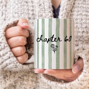 May include: White ceramic mug with vertical green and white stripes. The mug features the text "chapter 60" in black script, along with a small floral illustration. The mug is held by a person wearing a cream-colored sweater.