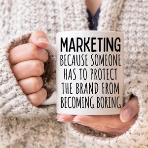 May include: White ceramic mug with black text that reads "MARKETING because someone has to protect the brand from becoming boring."