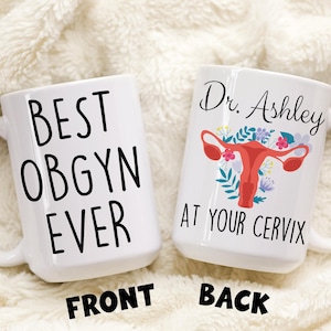 May include: White ceramic coffee mug with black text that reads "Best OBGYN Ever" on the front and "Dr. Ashley At Your Cervix" on the back. The back also features a pink uterus with flowers.