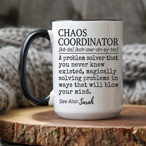 Puede incluir: Taza de cer&aacute;mica blanca con asa y borde negros. La taza presenta el texto "CHAOS COORDINATOR" con una definici&oacute;n y las palabras "See Also: Sarah". La taza est&aacute; sobre una superficie de madera.