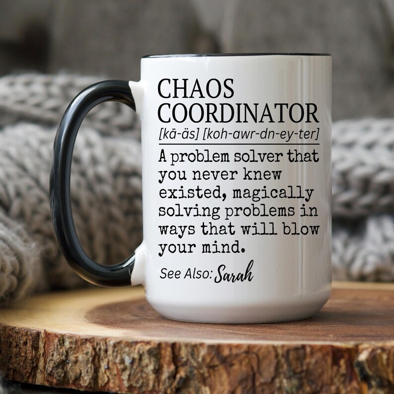 Puede incluir: Taza de cer&aacute;mica blanca con asa y borde negros. La taza presenta el texto "CHAOS COORDINATOR" con una definici&oacute;n y las palabras "See Also: Sarah". La taza est&aacute; sobre una superficie de madera.
