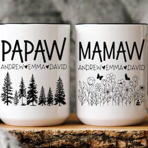 May include: Two white ceramic mugs with black handles. The mug on the left says "Papaw Andrew Emma David" with a forest scene. The mug on the right says "Mamaw Andrew Emma David" with a floral scene.
