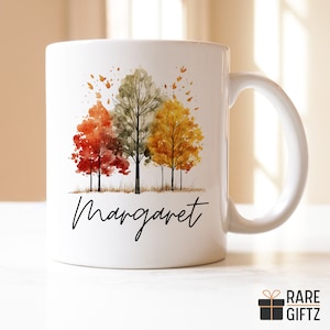 May include: White ceramic mug with a watercolor design of three trees with fall foliage in shades of red, green, and yellow. The mug has the name "Margaret" written in black cursive script.