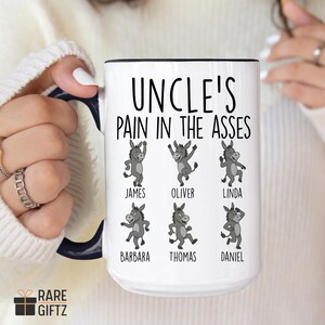 May include: White ceramic mug with a black rim and handle, featuring the text "UNCLE'S PAIN IN THE ASSES" in black. Below the text are six cartoon donkeys with names: James, Oliver, Linda, Barbara, Thomas, and Daniel. The mug is held by a person.