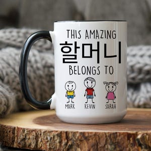 May include: White ceramic mug with a black handle. The mug has black text that reads "This amazing 할머니 belongs to" with three stick figures below the text labeled "Mark", "Kevin", and "Sarah".