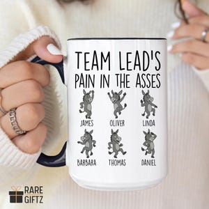 May include: White ceramic mug with a black rim and handle, featuring the text "TEAM LEAD'S PAIN IN THE ASSES" above cartoon donkeys. Each donkey has a name: James, Oliver, Linda, Barbara, Thomas, and Daniel. The mug is held by a person.
