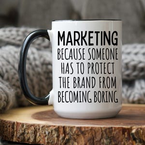 May include: White ceramic coffee mug with a black handle and black text that reads "Marketing because someone has to protect the brand from becoming boring."