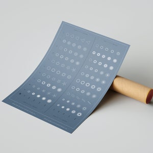 Pode incluir: Um poster azul-acinzentado com formas geométricas brancas e texto, parcialmente enrolado em torno de um tubo de cartão castanho. O poster apresenta vários desenhos circulares e em forma de estrela, com texto na parte superior e inferior. O tubo tem detalhes em vermelho.