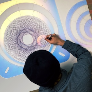 May include: A person is using a clear plastic stencil with concentric circles to create a complex geometric design with a pen. The stencil is on a white surface and has a blue, pink, and yellow gradient.