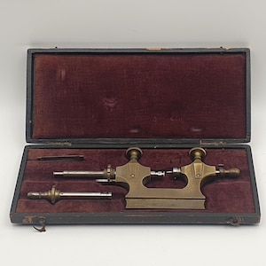 May include: An antique brass watchmaker's lathe tool set, presented in a black wooden case with a burgundy velvet lining. The set includes a lathe, a smaller tool, and additional components. The tools have a brass finish.