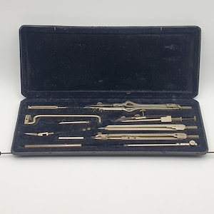Peut inclure: Un ensemble de dessin vintage dans un étui doublé de velours noir. L'ensemble comprend plusieurs compas, diviseurs et autres outils de dessin argentés. L'étui est ouvert, révélant les outils soigneusement rangés à l'intérieur.