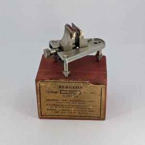 May include: A silver-toned metal Bergeon balance poising tool with two upright jaws and a leveling base, mounted on a red wooden box. The box features a tan label with the text BERGEON, No. 3055, and descriptions in French, German, and English.