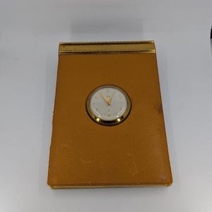Pode incluir: Um acessório de secretária retangular revestido a couro castanho-claro, com uma borda superior em metal dourado e um relógio analógico circular centrado com mostrador branco, ponteiros e numerais dourados numa moldura de metal dourado polido.