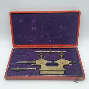 Puede incluir: Un juego de herramientas de torno para relojero de latón en una caja rectangular de madera roja con interior de terciopelo morado. El set incluye un accesorio de torno central de latón con dos perillas de ajuste, dos husillos de metal y un pasador.