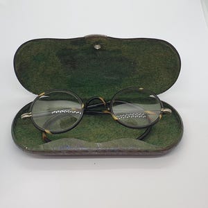 Könnte beinhalten: Runde Brille mit Schildpattmuster, offen in einem grünen Etui. Die Brille hat einen dünnen, schwarzen Rahmen und klare Gläser. Das Etui ist oval und hat einen Knopfverschluss.