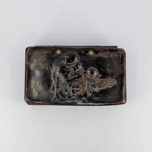 Puede incluir: Una caja rectangular de madera de color marrón oscuro con un diseño en relieve tallado en la tapa. La talla representa una figura en movimiento con líneas fluidas y sutiles acentos dorados integrados en la textura de la madera oscura.