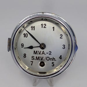 May include: A round silver-toned clock with a metallic face featuring black Arabic numerals from 1 to 12. The center displays the text M.V.A.-2 and S.M.V./Onh. in black lettering. It has two black hands and a polished chrome-colored circular frame.