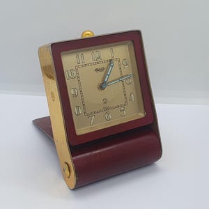 May include: Vintage Jaeger travel alarm clock with a burgundy leather case and gold-tone accents. The clock face is a light beige with black numerals and blue hands. The clock is rectangular and folds for storage.