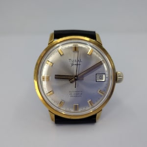 Peut inclure: Une montre-bracelet automatique Tusal Geneve avec un boîtier rond doré, un cadran argenté à effet soleil et un bracelet en cuir noir. Le cadran présente des index dorés, un guichet de date à trois heures et l'inscription Tusal Geneve.