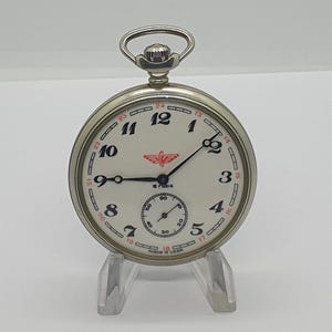 May include: A silver-toned pocket watch with a white dial, black Arabic numerals, and a red winged logo. The watch features a sub-dial for seconds, black hands, and the text 18 Rubis and MADE IN USSR printed on the face. It has a top winding crown.
