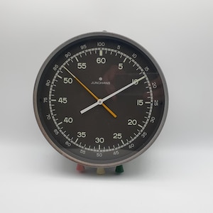 May include: A vintage Junghans stopwatch with a black face, white numerals, and a silver-tone bezel. The clock features white and orange hands and three colored buttons at the bottom. The clock measures approximately 4 inches in diameter.