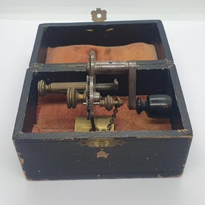 Könnte beinhalten: Antiker Musikbox-Mechanismus in einem schwarzen Holzkasten. Der Mechanismus besteht aus Messing- und Stahlkomponenten, darunter Zahnräder, eine Kurbel und eine kleine Kette. Das Innere des Gehäuses ist mit orangefarbenem Stoff ausgekleidet.