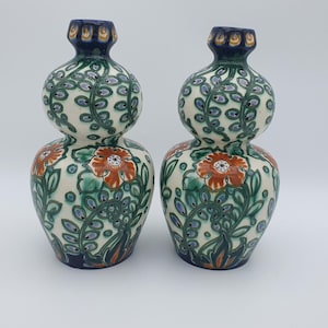 May include: Two ceramic vases with a double-bulb shape. The vases feature a cream-colored background with a floral pattern of orange flowers, green leaves, and blue accents. The rims and bases are dark blue.