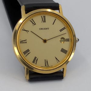 Puede incluir: Un reloj de pulsera Orient con caja redonda en tono dorado y esfera color crema con números romanos negros. El reloj incluye manecillas negras delgadas, una ventana de fecha en la posición de las tres y una correa de cuero negro.