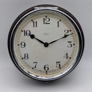 Könnte beinhalten: Eine runde analoge Wanduhr mit dunkelbraunem Rahmen und verchromter Lünette. Das cremefarbene Zifferblatt zeigt schwarze arabische Ziffern, schwarze Zeiger und den Schriftzug "SMITHS 7 JEWELS EIGHT DAY" oben sowie "MADE IN GREAT BRITAIN" unten.