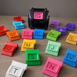 May include: A black plastic letter dispenser unit with a pink G tile inserted, surrounded by various colorful plastic letter tiles in red, orange, yellow, green, blue, purple, pink, and white, each featuring a single black uppercase letter.