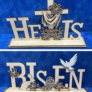 May include: Wooden Easter decorations with the phrases "He Is Risen". The top piece features a cross and floral accents. The bottom piece includes a bunny, flowers, a basket, and a dove. The decorations are on a wooden base.