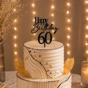 Könnte beinhalten: Eine zweistöckige weiße Torte mit texturierten horizontalen Rillen und Blattgold-Akzenten, gekrönt von einem schwarzen Acryl-Tortenstecker mit der Aufschrift Happy Birthday 60 in Schreib- und Serifenschrift sowie filigranen goldenen Blattverzierungen.
