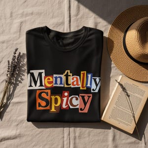 May include: Black t-shirt with the text "Mentally Spicy" in a colorful, cut-out letter style. The shirt is folded on a beige surface, with a straw hat and an open book nearby. A sprig of lavender is also present.