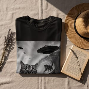 May include: Black t-shirt featuring a grayscale graphic of cats and UFOs. One cat looks surprised, while another surfs a wave created by a UFO. Several flying saucers are in the sky, creating a unique and humorous design.