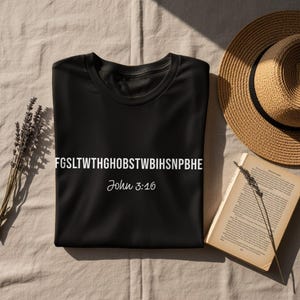 May include: A black t-shirt with white text reading "FGSLTWTHGHOBSTWBIIHSNPBHE" and "John 3:16". The shirt is on a beige surface, with a straw hat and an open book nearby.