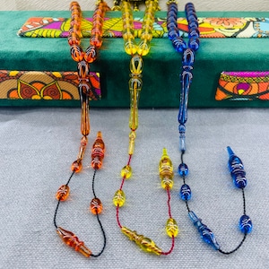 May include: Three prayer beads in amber, yellow, and blue. Each strand features a series of rounded beads and a decorative pendant. The beads are strung on black and red cords. The background includes a green velvet surface and a patterned fabric.