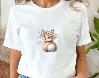 Puede incluir: Camiseta blanca con un gráfico de un conejito marrón que lleva una corona de flores rosas. El texto "SPRING is in the air" está impreso debajo del conejito.
