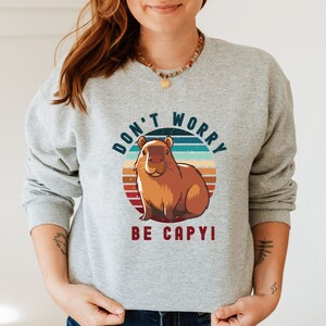 Op de afbeelding: Een grijs sweatshirt met een vintage-stijl grafische afbeelding van een capibara in een cirkel met de tekst "Don't Worry Be Capy!" gedrukt op de voorkant.