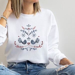Op de afbeelding: Witte sweatshirt met een folk art design met twee blauwe vogels, rode harten en bloemelementen. Het ontwerp is gecentreerd op de borst. De sweatshirt heeft lange mouwen en een ronde hals.