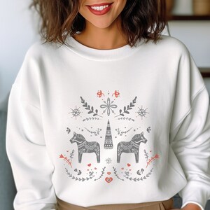 Puede incluir: Sudadera blanca con un diseño de arte popular escandinavo. El diseño incluye dos caballos Dala grises, elementos florales, copos de nieve y una torre central, todo en acentos grises y rojos. Una prenda superior cómoda y elegante.