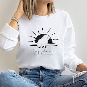 Op de afbeelding: Wit sweatshirt met een zwart-witte illustratie van een capybara voor een zonsondergang. De tekst "Capybara is my spiritual animal" is onder de illustratie gedrukt.