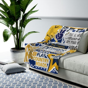 May include: A yellow, blue, and white throw blanket with text that reads "West Virginia", "Country Roads", and "This is what dreams are made of". The blanket is draped over a white couch.