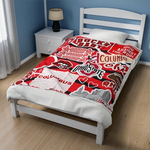 Puede incluir: Una manta de tiro roja y negra con un collage de gráficos temáticos de los Ohio State Buckeyes, incluyendo las palabras "Ohio State", "Buckeyes", "Columbus" y "Ohio Against the World".