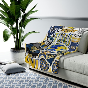 May include: A white, blue, and yellow fleece blanket draped over a gray sofa. The blanket features a collage of graphics and text, including "TCNJ", stars, and a lion. The blanket is a cozy home decor item.