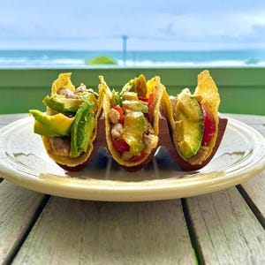 Op de afbeelding: Drie taco's gevuld met avocado, tomaten en andere ingrediënten, gepresenteerd op een wit bord. De taco's worden in een houten houder gehouden. De achtergrond toont een uitzicht op de oceaan en de lucht.