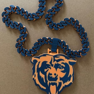 May include: A necklace featuring a thick, chunky chain with alternating dark blue and orange links. A large, flat pendant shaped like a roaring bear head in dark blue and orange hangs from the centre of the chain, attached by a small blue loop.