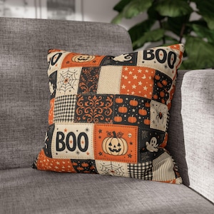 Pode incluir: Uma almofada quadrada com um design de patchwork nas cores laranja, preto e creme. O padrão inclui ilustrações de abóboras, fantasmas, aranhas, teias de aranha, estrelas e o texto BOO impresso em letras maiúsculas pretas e negrito.