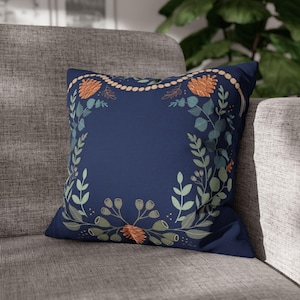 Pode incluir: Uma almofada quadrada azul-marinho com um design central de coroa botânica. A estampa inclui folhas de eucalipto verde-claro, pequenas vagens cinzentas e pinhas cor de laranja ligadas por um fio de contas de madeira clara.