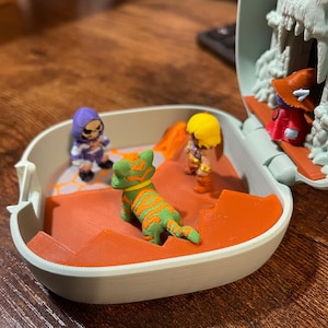 Op de afbeelding: Een kleine speelset met verschillende miniatuurfiguren. De figuren omvatten een groen-oranje tijger, een paars-wit skelet en twee andere personages in gele en rode kleding. De speelset heeft een lichtblauwe buitenkant en een oranje binnenkant.
