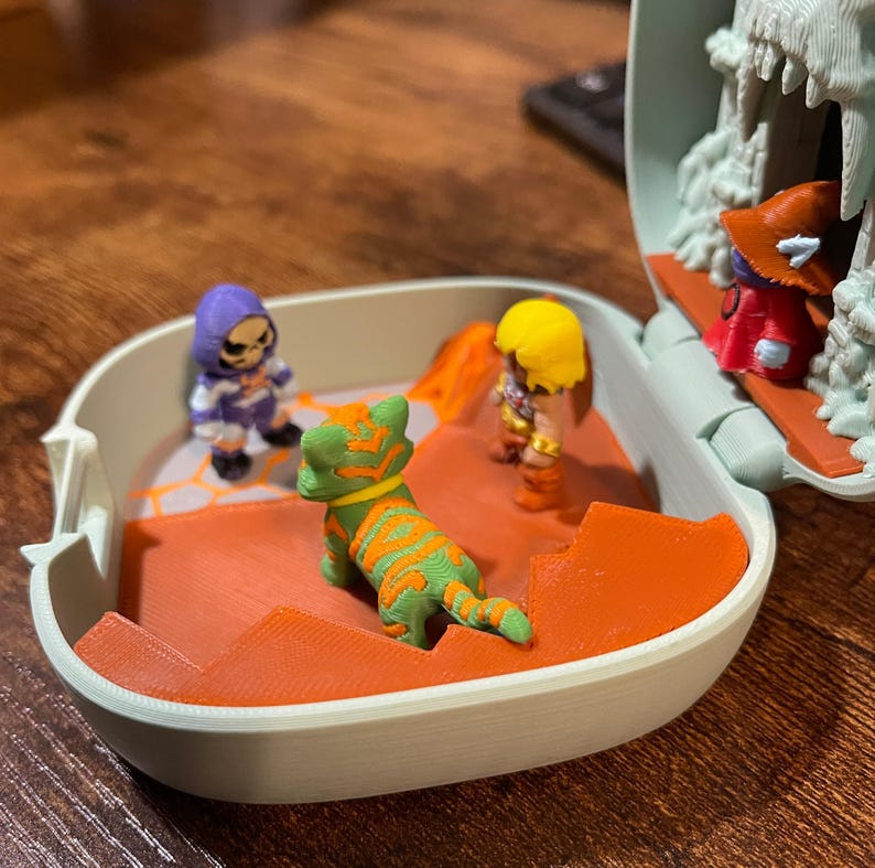 Op de afbeelding: Een kleine speelset met verschillende miniatuurfiguren. De figuren omvatten een groen-oranje tijger, een paars-wit skelet en twee andere personages in gele en rode kleding. De speelset heeft een lichtblauwe buitenkant en een oranje binnenkant.